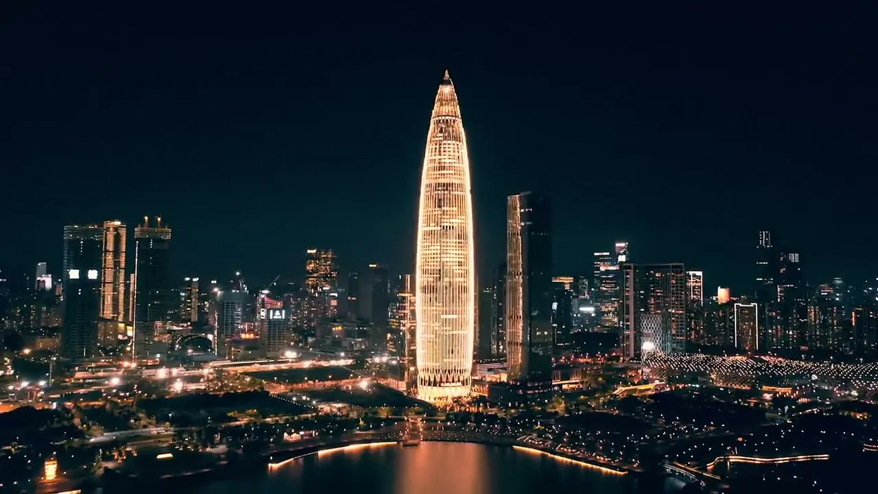 Aerial view of the Shenzhen skyline at night, a floodlit tower rising above the waterfront